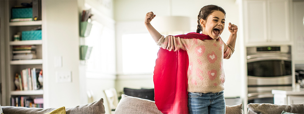 Ein Mädchen trägt einen Superheldinnen Umhang und steht in einer Power-Pose auf dem Sofa im Wohnzimmer