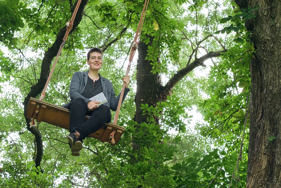 Liv Schlett, die kreatives Schreiben studiert hat, sitzt auf einer Schaukel am Baum, über ihr die Baumwipfel und in der Hand hält sie ein Buch und ist glücklich, weil sie die Angst vor Menschen verloren hat.