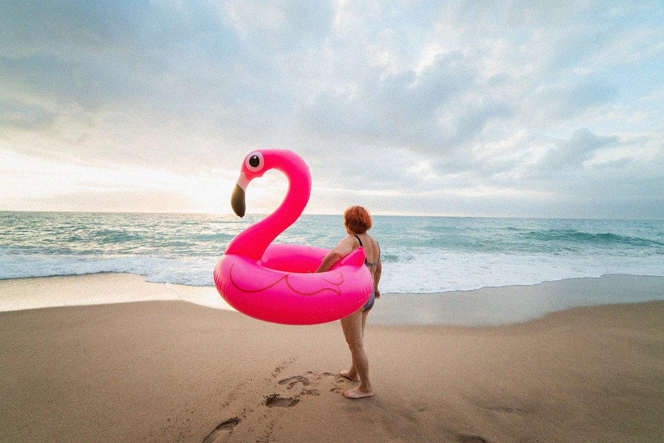 Eine ältere Frau mit rötlichen Haaren steht mit einem aufblasbaren Flamingo an einem einsamen Strand