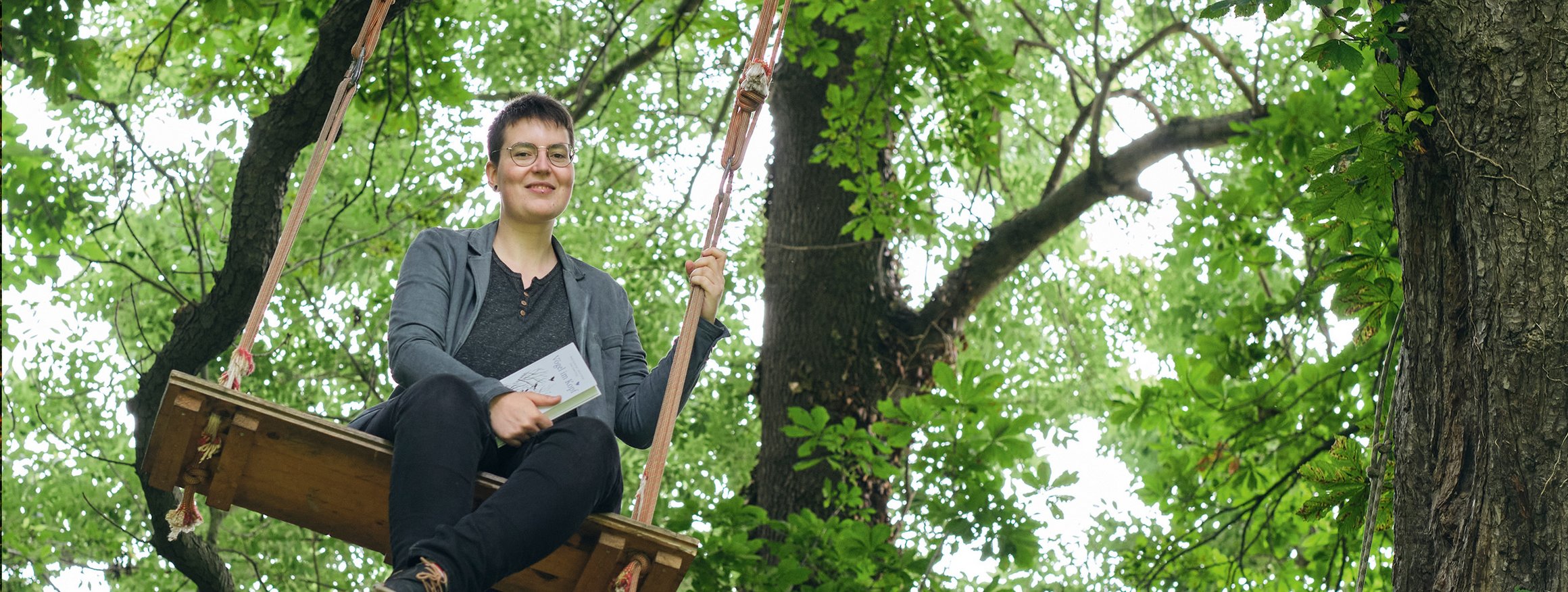 Liv Schlett, die kreatives Schreiben studiert hat, sitzt auf einer Schaukel am Baum, über ihr die Baumwipfel und in der Hand hält sie ein Buch und ist glücklich, weil sie die Angst vor Menschen verloren hat.