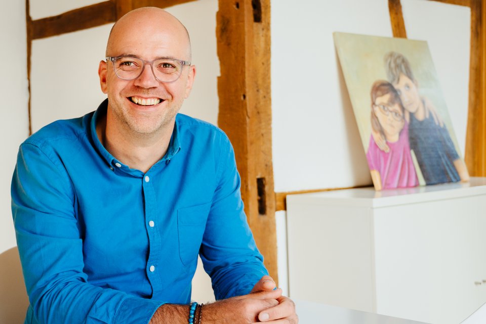 Ein Mann mit Brille und blauem Hemd sitzt lachend auf einem Stuhl, die Hände gefaltet und hinter ihm an die Wand gelehnt, steht das Porträt seiner Kinder