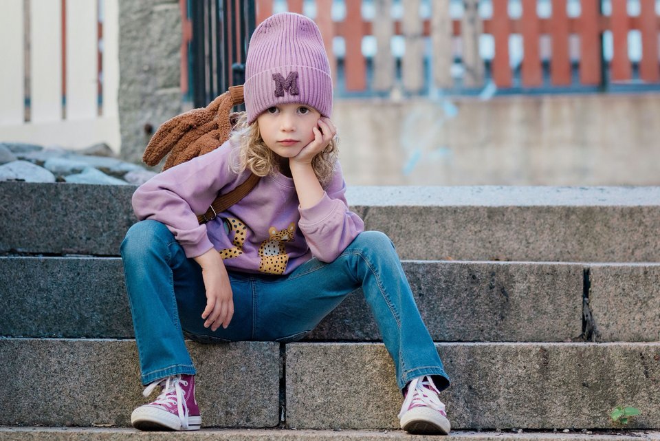 Ein kleines Mädchen sitzt nachdenklich mit Wollmütze und und Jeans auf einer Treppe und hat das Kinn auf die Hand gestützt