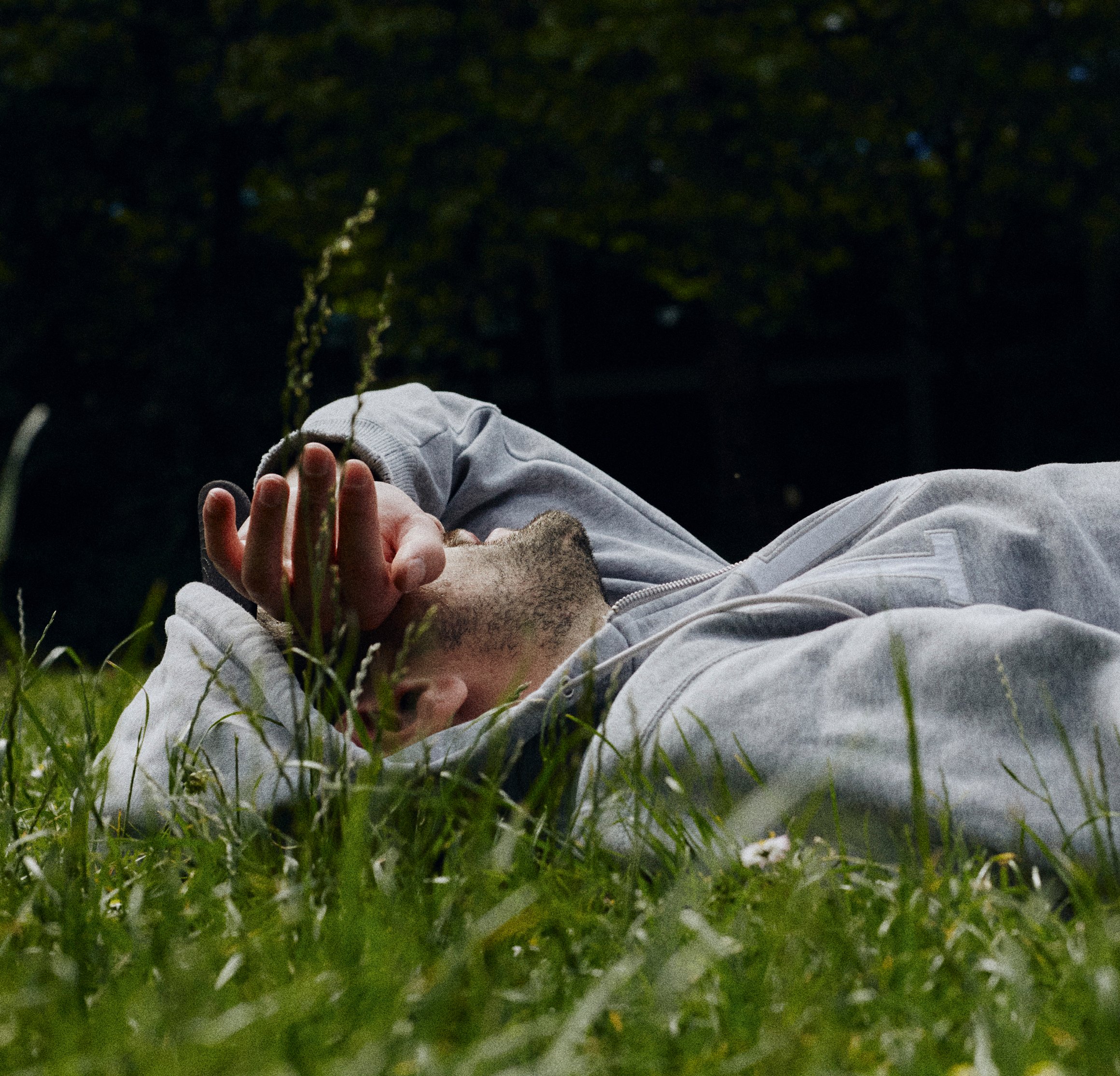 Das Foto zeigt einen Mann in Kapuzenpulli, der mit dem Rücken auf einer Wiese liegt und mit seinem Handrücken die Augen verdeckt.