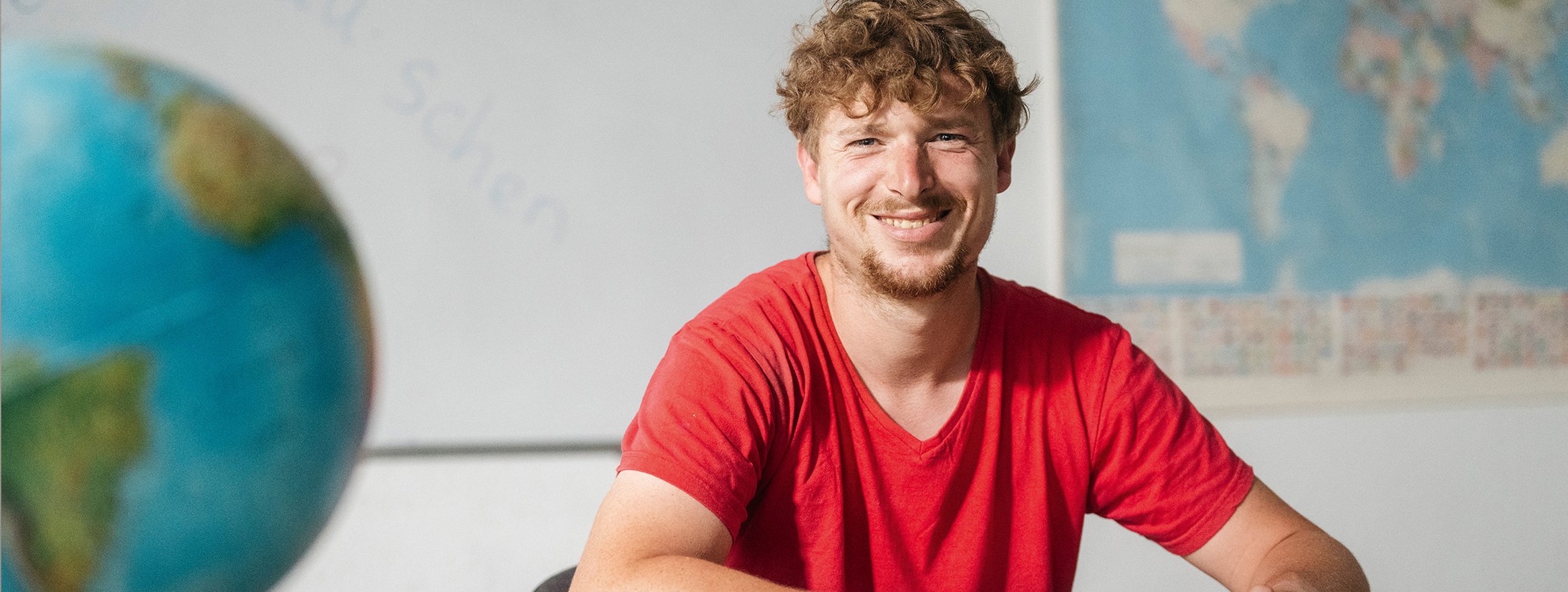 Ein junger Mann im roten T-Shirt sitzt lächelnd in einem Grundbildungskurs der Volkshochschule am Schreibtisch und verbessert sein Lesen 