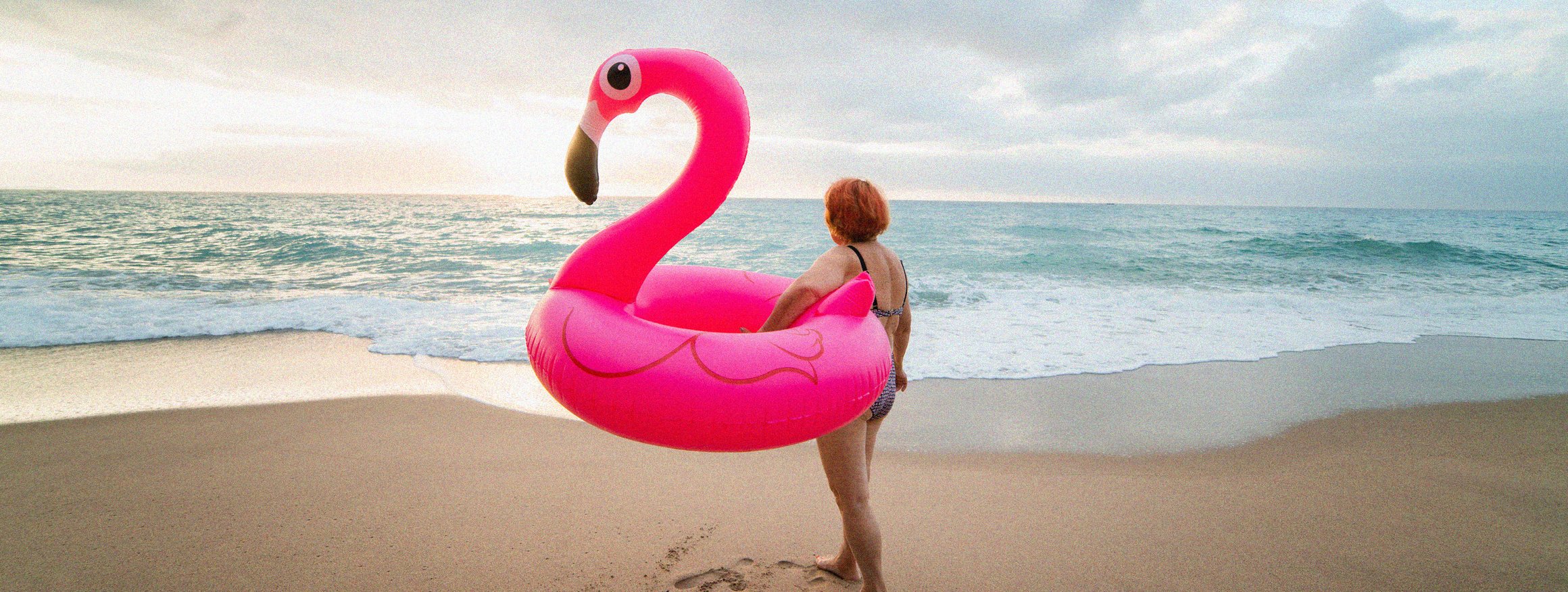 Eine ältere Frau mit rötlichen Haaren steht mit einem aufblasbaren Flamingo an einem einsamen Strand