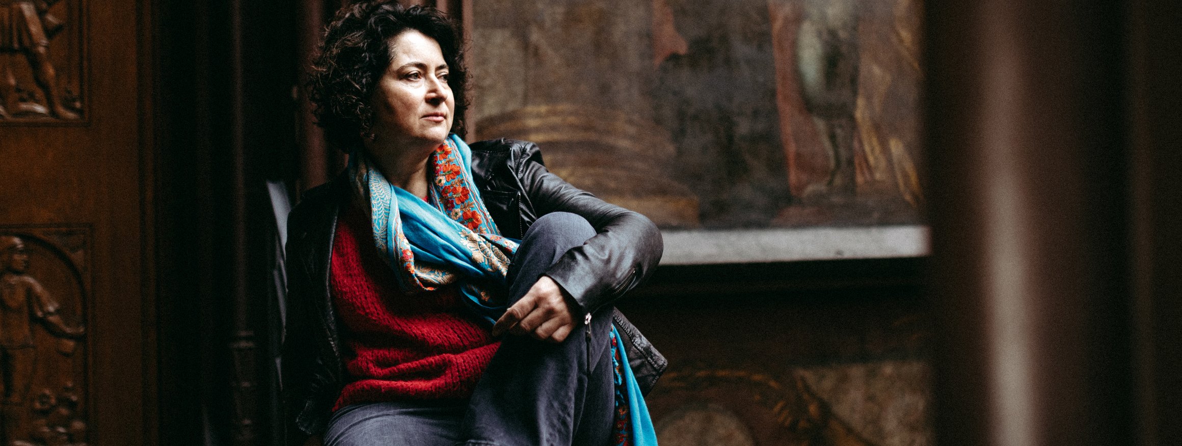 Eine Frau mit dunklen Locken, Jeans und Lederjacke sitzt mit angewinkeltem Bein da und schaut ernst aus dem Fenster