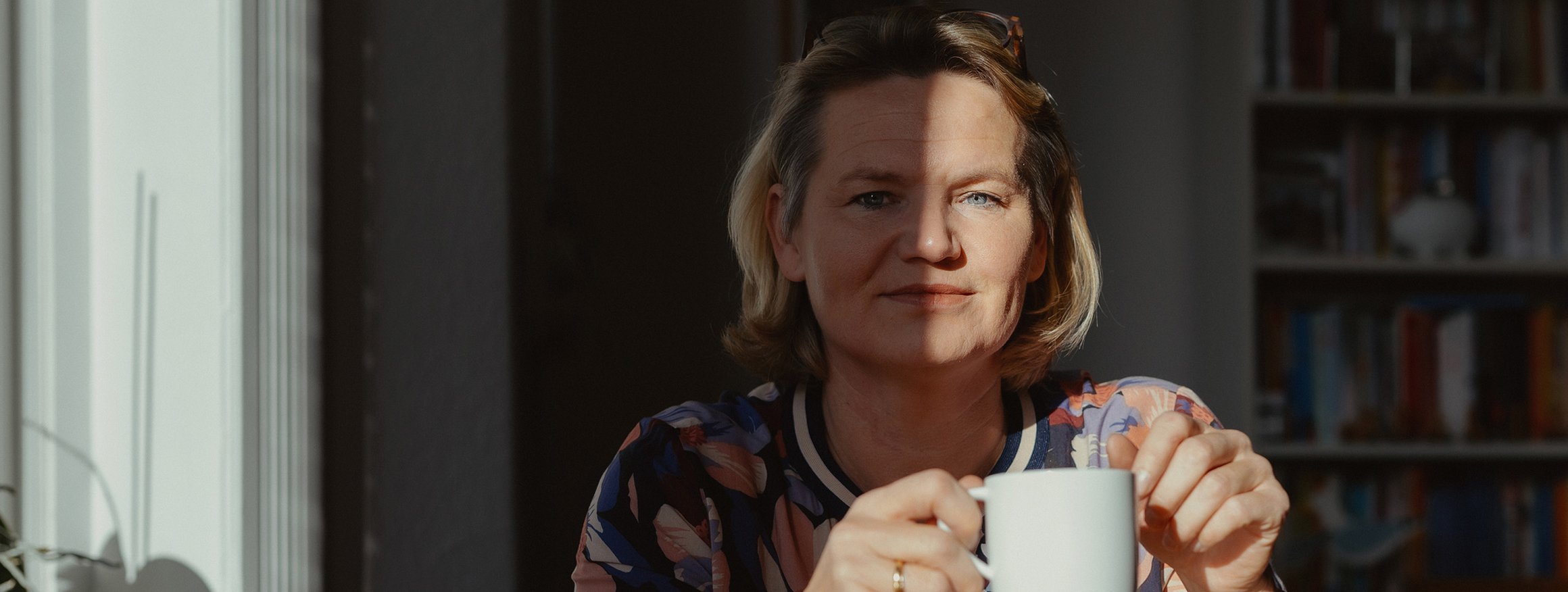 Sandra Schumacher sitzt im Halbschatten an einem Tisch und hält eine weiße Tasse in der Hand und ist froh, dass sie seit 20 Jahren ohne Alkohol lebt