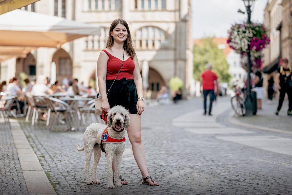 Eine junge Frau steht lächelnd und selbstsicher in einer Altstadtkulisse mit ihrer Labradoodle-Hündin, die noch in Ausbildung zur Assistenzhündin ist