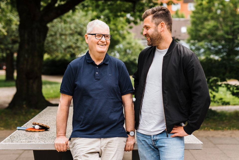 Das Foto zeigt Vater und Sohn, die an einer Tischtennisplatte gelehnt miteinander lachen.