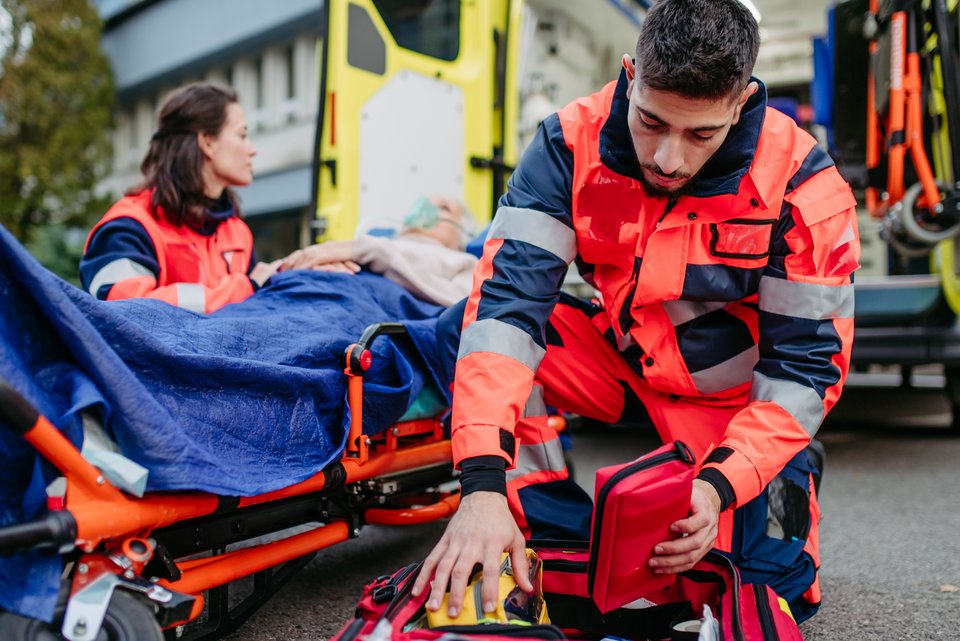Eine Sanitäterin und ein Sanitäter versorgen eine verletzte Person auf einer Liege, während dahinter der Krankenwagen steht, bereit zum Krankentransport