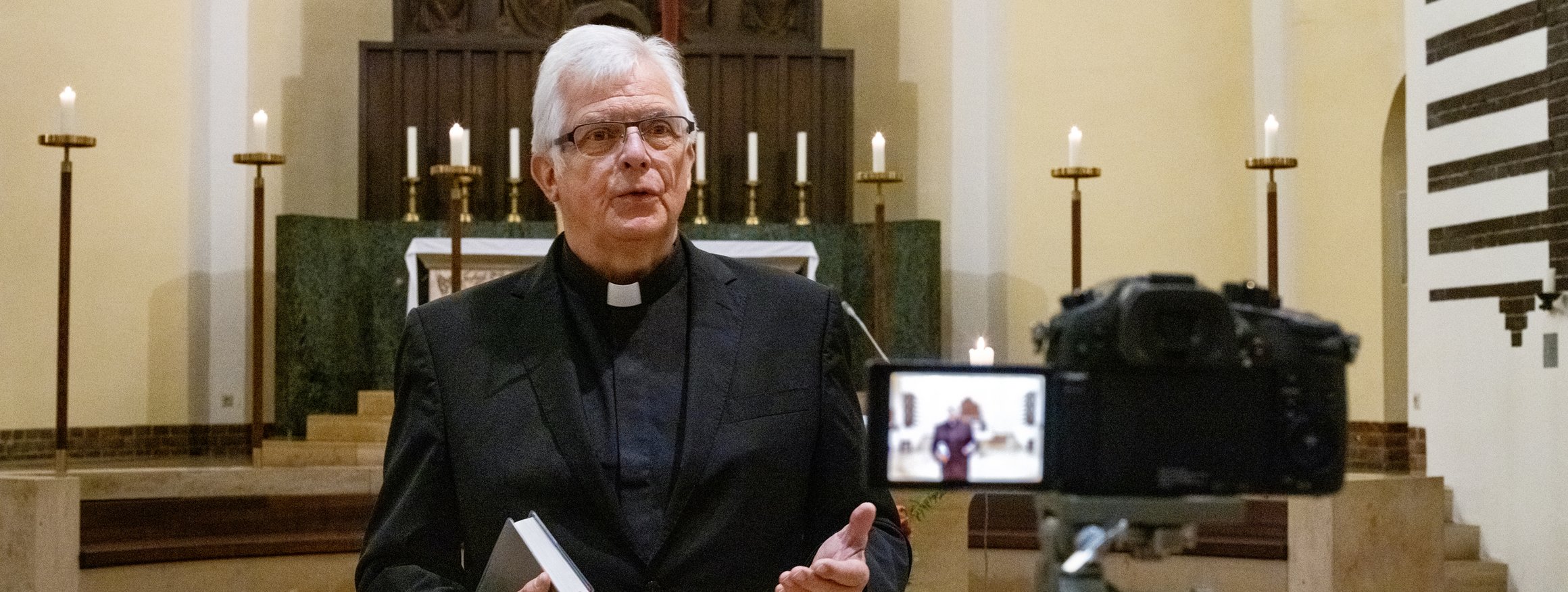 Ein Pfarrer steht vor dem Altar in einer Kirche und spricht und wird dabei gefilmt