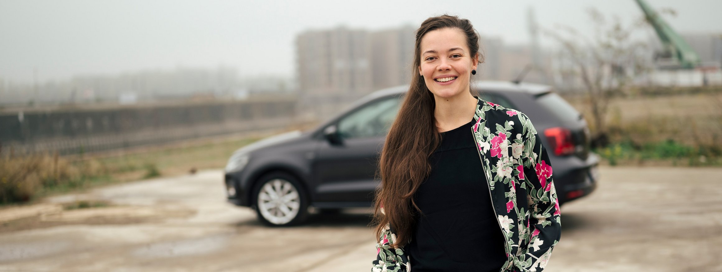 Eine junge Frau mit langen dunklen Haaren, steht lächelnd mit den Händen in den Taschen ihrer schwarzen Jeans, vor einem Auto und hat die Angst vor dem Autofahren überwunden