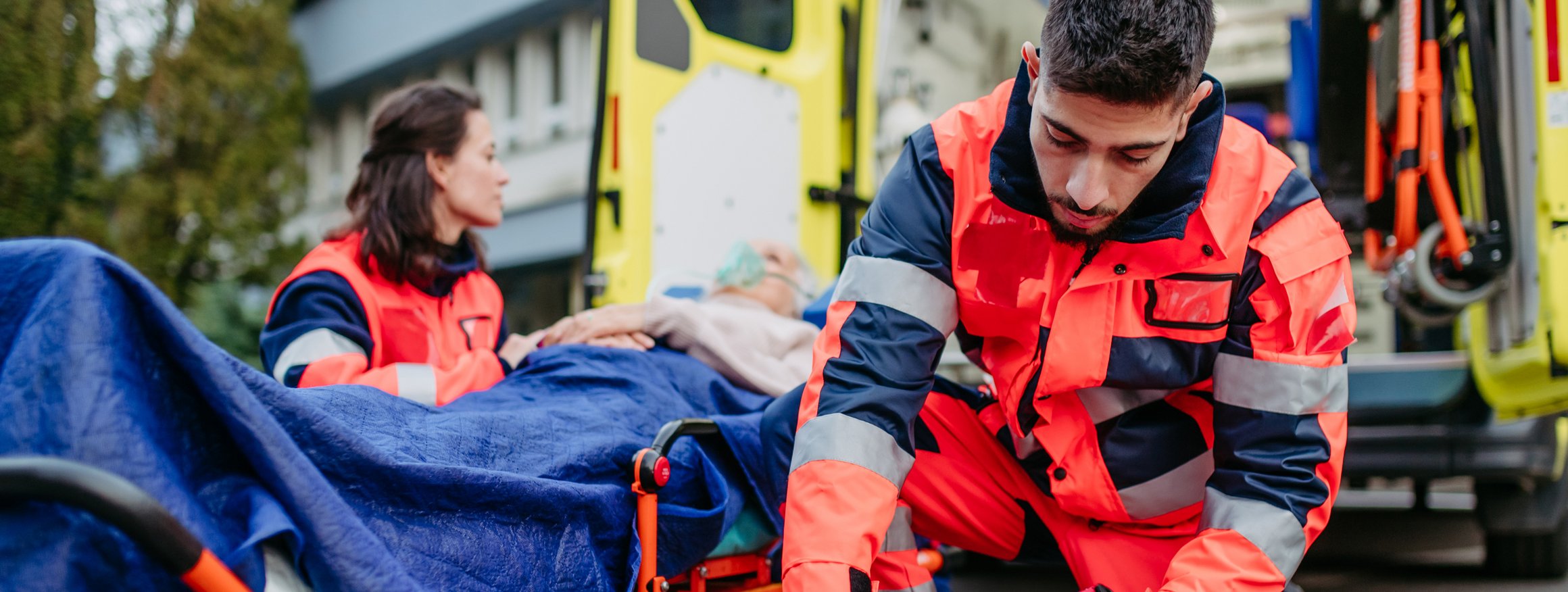 Eine Sanitäterin und ein Sanitäter versorgen eine verletzte Person auf einer Liege, während dahinter der Krankenwagen steht, bereit zum Krankentransport