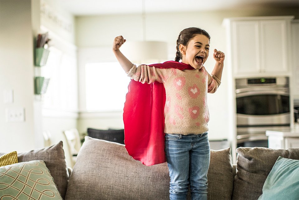 Ein Mädchen trägt einen Superheldinnen Umhang und steht in einer Power-Pose auf dem Sofa im Wohnzimmer