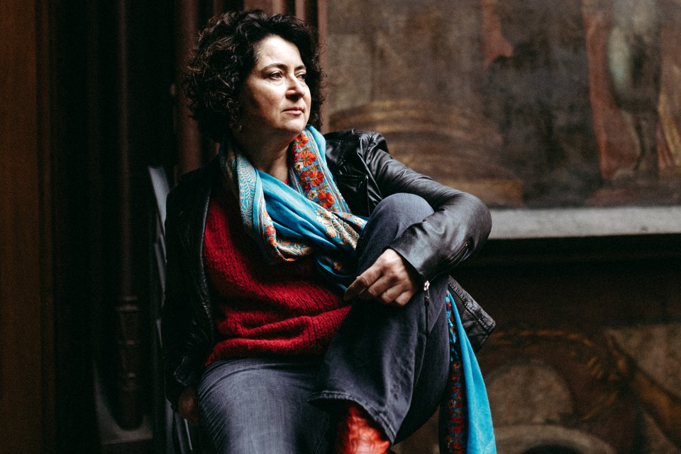 Eine Frau mit dunklen Locken, Jeans und Lederjacke sitzt mit angewinkeltem Bein da und schaut ernst aus dem Fenster