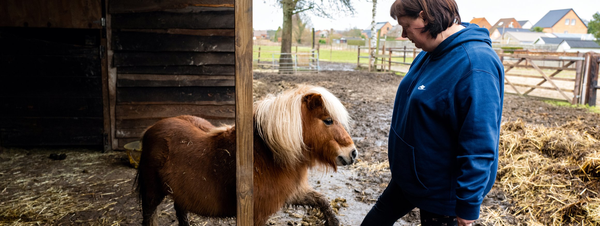 Eine Frau steht mit Stiefeln im Stall und kümmert sich um ein Pony, wobei beide spielerisch ein Bein heben