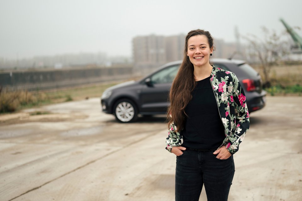 Eine junge Frau mit langen dunklen Haaren, steht lächelnd mit den Händen in den Taschen ihrer schwarzen Jeans, vor einem Auto und hat die Angst vor dem Autofahren überwunden