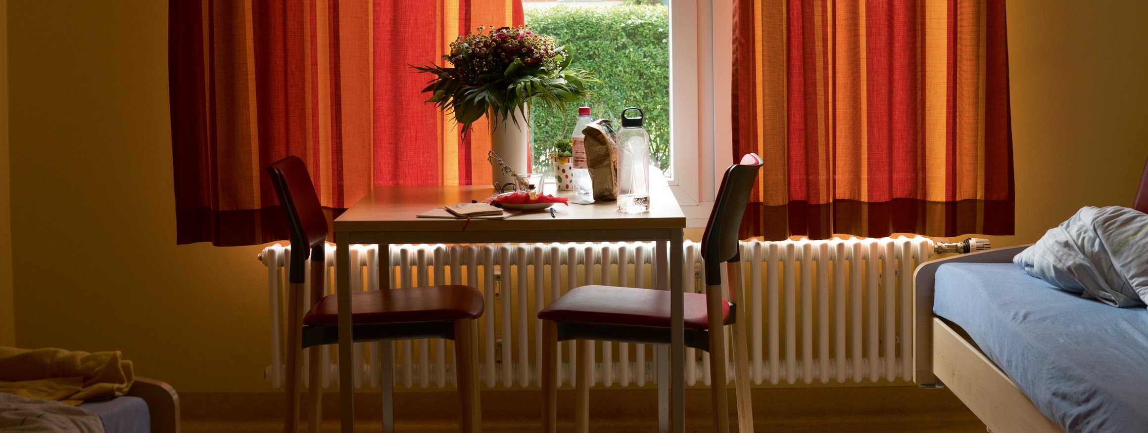 Ein Zimmer in der Psychiatrie mit Bett, Tisch, zwei Stühlen und ein Blumenstrauß steht auf dem Tisch, während die Vorhänge am Fenster halb zugezogen sind