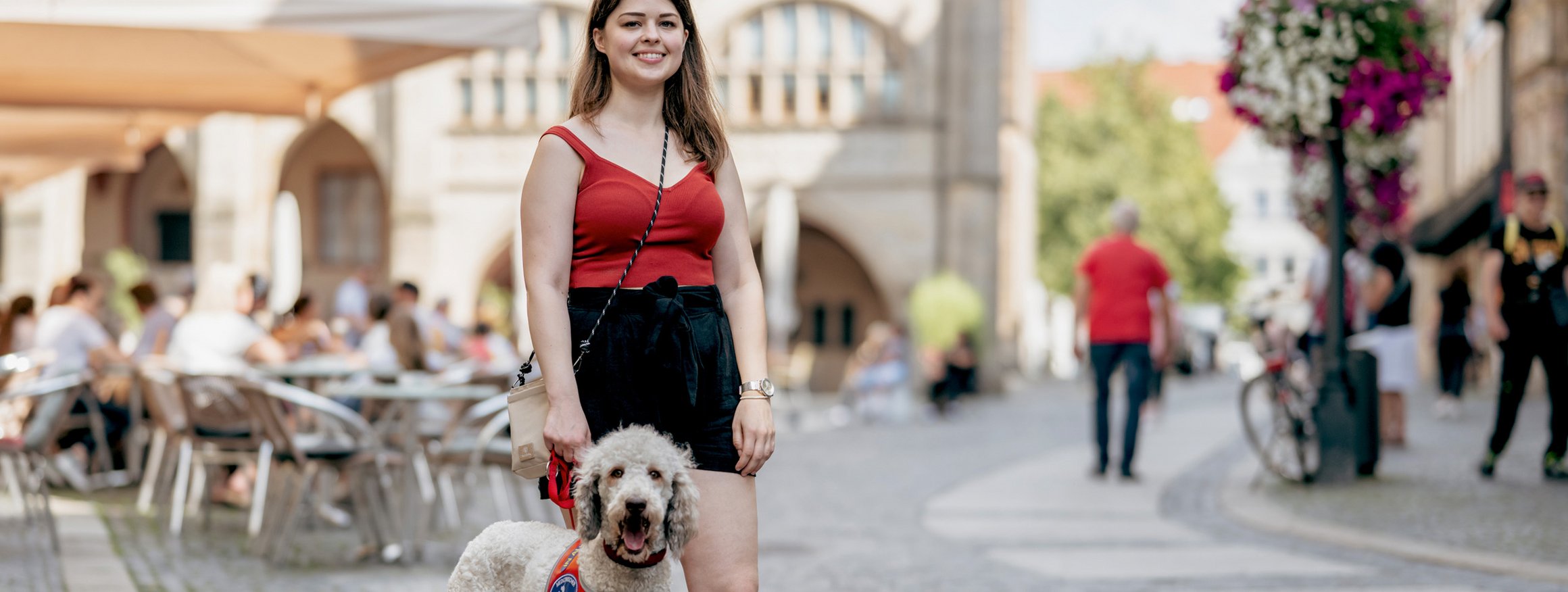 Eine junge Frau steht lächelnd und selbstsicher in einer Altstadtkulisse mit ihrer Labradoodle-Hündin, die noch in Ausbildung zur Assistenzhündin ist