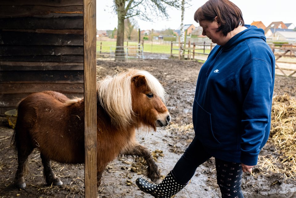 Eine Frau steht mit Stiefeln im Stall und kümmert sich um ein Pony, wobei beide spielerisch ein Bein heben