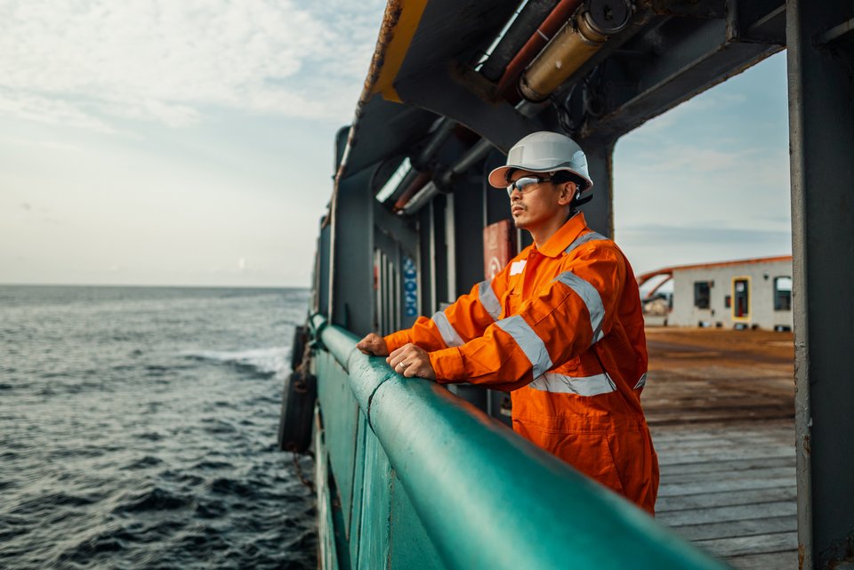 Ein Arbeiter mit Helm und Arbeitskleidung steht an der Reling auf einem Schiff und schaut auf das Meer