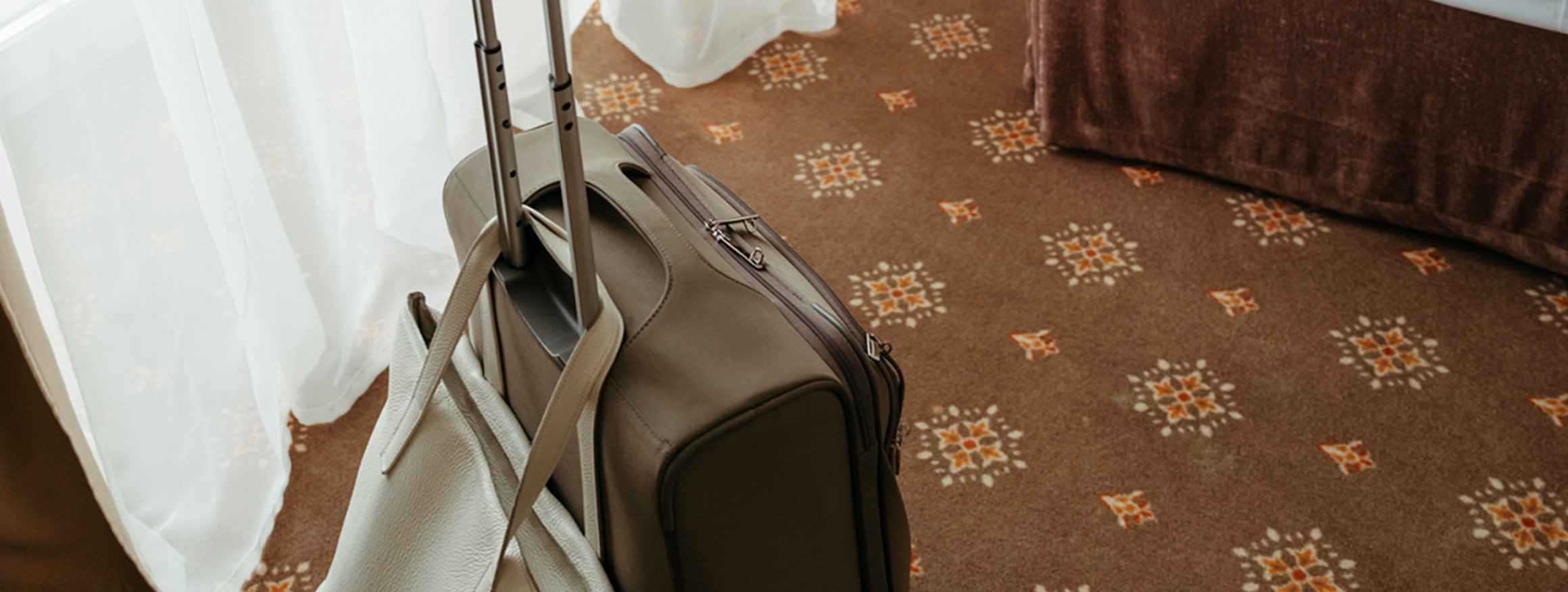 Ein Rollkoffer, an dem eine Handtasche hängt, steht auf dem Teppich eines Hotelzimmers