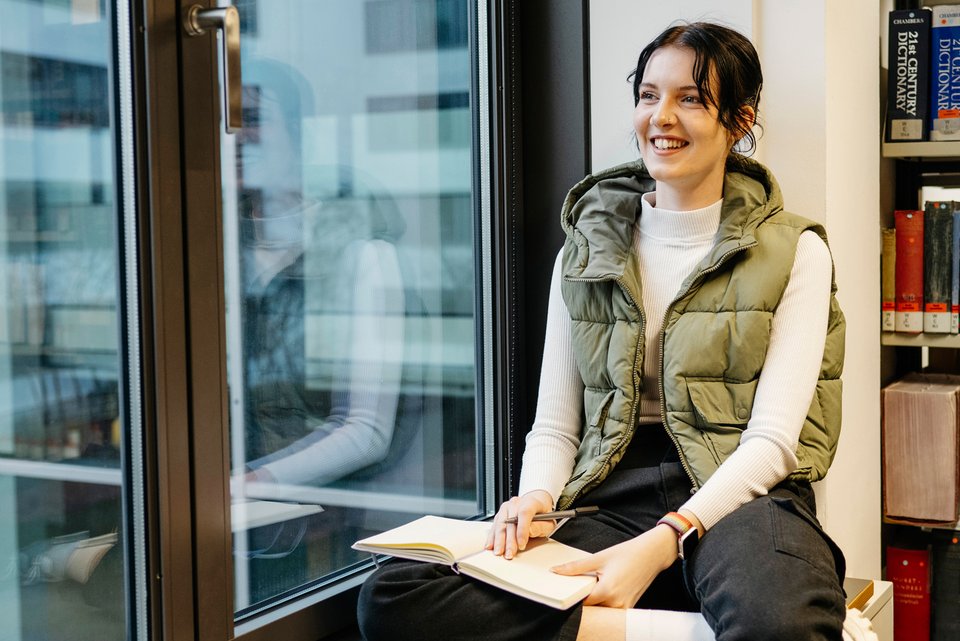 Eine junge Frau sitzt in einem Arbeitszimmer lächelnd am Fenster und schreibt in ein Buch