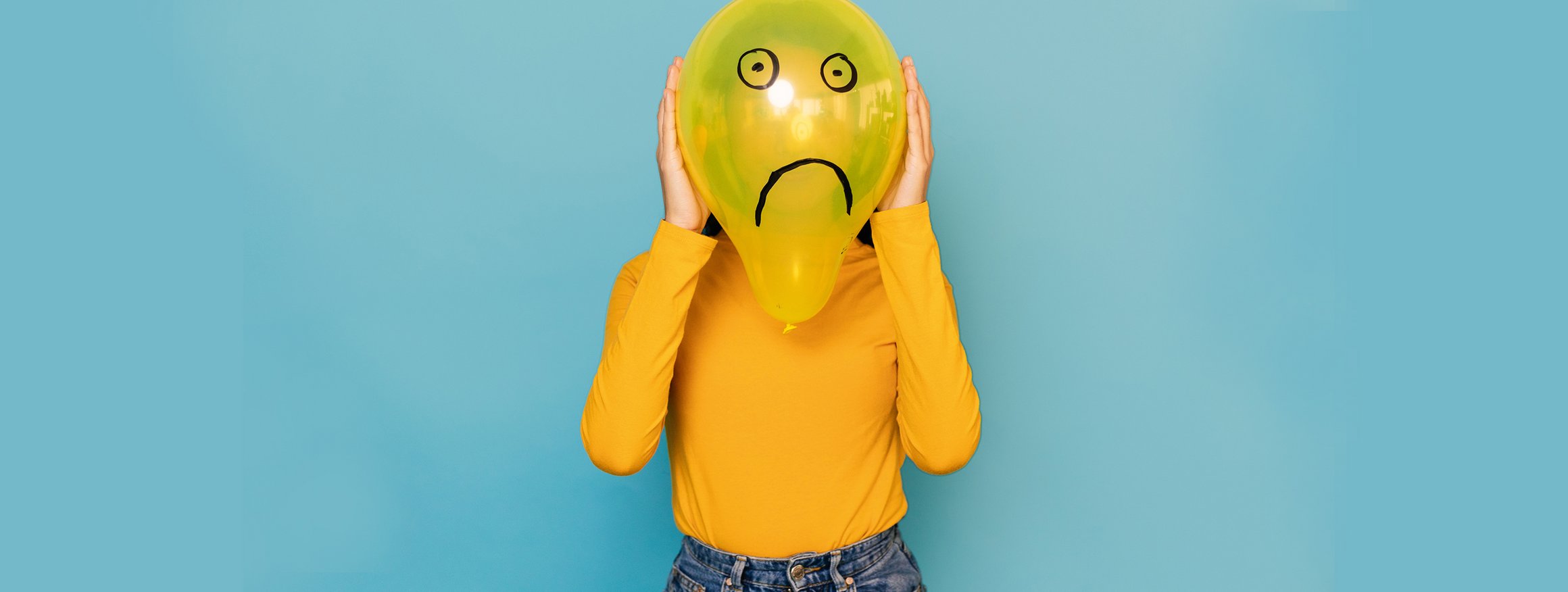 Eine junge Frau mit gelben Pulli hält einen gelben Luftballon vor ihr Gesicht, auf dem mit Filzstift, Augen und ein trauriger Mund gemalt sind