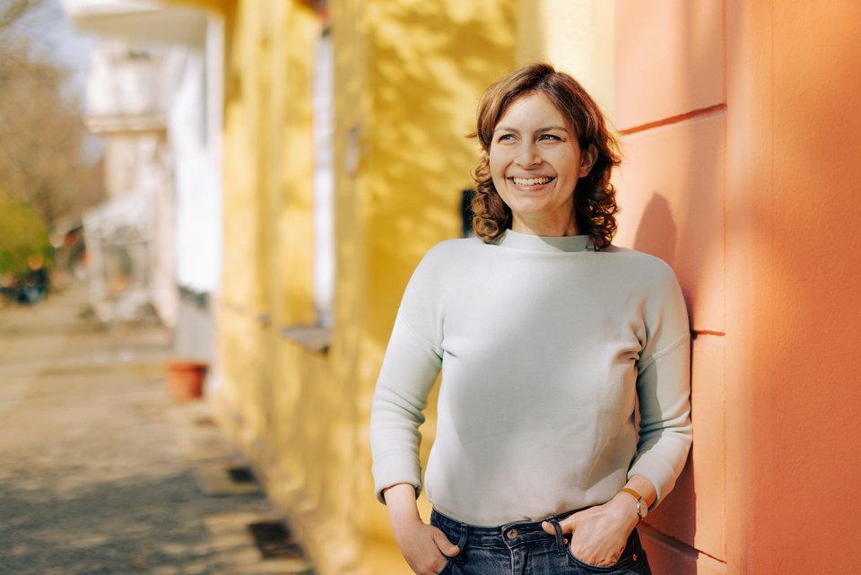 Eine junge Frau lehnt lächelnd an einer Hauswand, die Hände lässig in den Jeanstaschen und hat keine Angst mehr aufzufallen