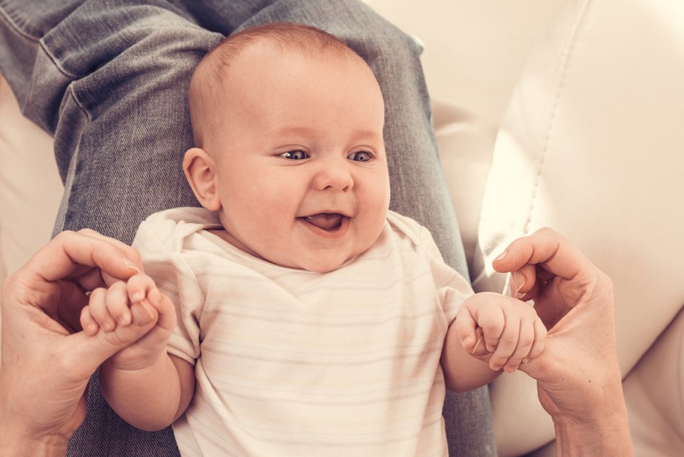 Eine Person hat ein Baby auf dem Schoß liegen und hält dabei die kleinen Händchen, während das Baby fröhlich ist
