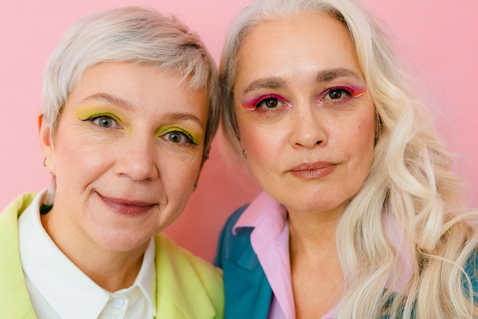 Zwei ältere Frauen in farbenfroher Kleidung und farbigen Augen-Makup, stehen nah beieinander