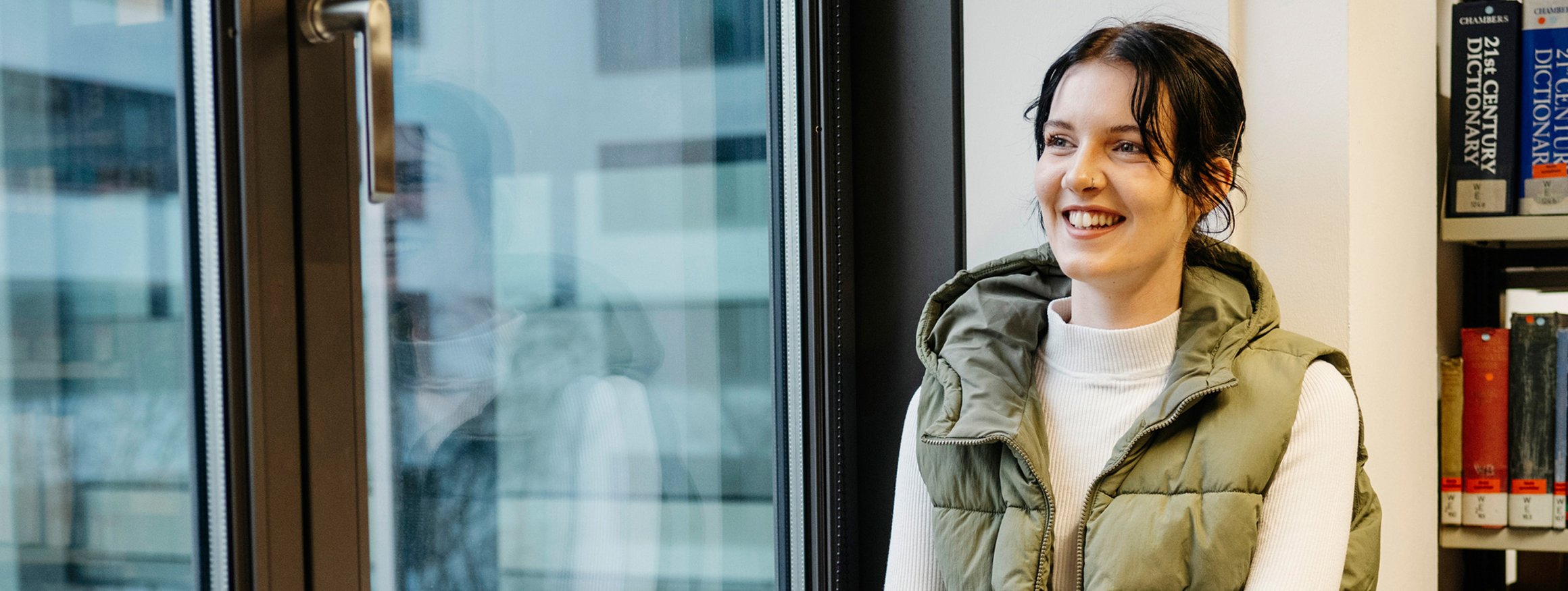 Eine junge Frau sitzt in einem Arbeitszimmer lächelnd am Fenster und schreibt in ein Buch